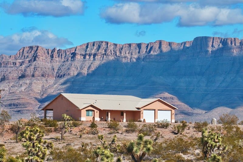 A single-story stucco home with a tile roof in the desert, red-rock mountains behind it