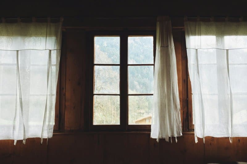 A wood-framed window with sheer curtains in a timber-walled room, hills beyond
